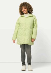 Chaqueta acolchada verde con un patrón de corazones texturizado, cierre frontal y capucha. Combinada con jeans grises y zapatillas blancas.