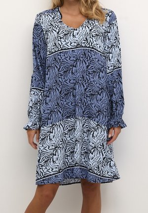 Kobieta w sukience za kolano z długim rękawem, ozdobionej niebiesko-czarnymi abstrakcyjnymi wzorami zebry i falbanami na mankietach.