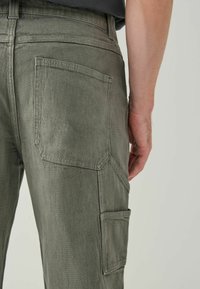 Green cargo pants featuring a textured fabric, multiple large pockets, and visible stitching details. Close-up of the back view.
