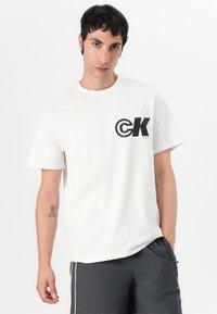 Balta medvilninė marškinėlių su juodu „CK“ logotipu kairėje krūtinės pusėje. Dizainas apima apvalią apykaklę ir trumpas rankoves.