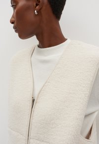 Fleeceweste in Creme mit Reißverschluss, weichem Stoff und V-Ausschnitt. Getragen über einem hellen, gerippten weißen Oberteil.