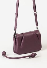 Piccolo borsa a tracolla in pelle viola con finitura a ciottoli, dotata di spallaccio regolabile e manico annodato amovibile, su uno sfondo bianco.