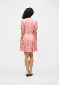 Γυναίκα με σγουρά μαλλιά που φοράει ένα ροζ φόρεμα με λουλουδάτο μοτίβο και φουσκωτά μανίκια, μαζί με μαύρα mules με τακούνι, γυρισμένη πλάτη σε απλό φόντο.