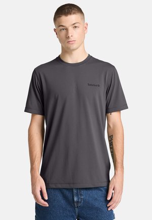 Młody mężczyzna stoi, ubrany w ciemnoszarą koszulkę Timberland i niebieskie dżinsy, z widocznym tatuażem na lewym przedramieniu.