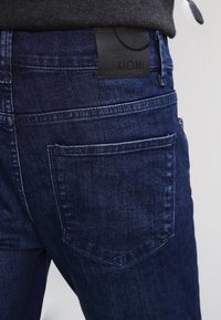 Dark blue denim jeans featuring a textured finish, with a black leather patch at the back displaying the brand "KIOMI."