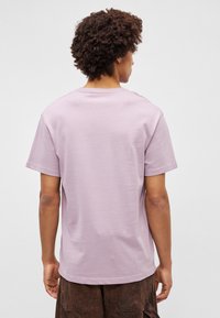 Λιλά βαμβακερό t-shirt με κοντά μανίκια και στρογγυλή λαιμόκοψη, φαίνεται από πίσω. Λείος υφασμάτινος σχεδιασμός, χαλαρή εφαρμογή, το τελείωμα επεκτείνεται ελαφρώς κάτω από τη μέση.