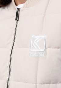 Beżowa pikowana kurtka z czarnym zamkiem i białym wyszywanym logo "KANI" na piersi.
