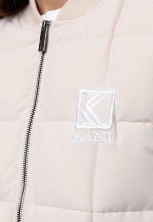 Beżowa pikowana kurtka z czarnym zamkiem i białym wyszywanym logo "KANI" na piersi.