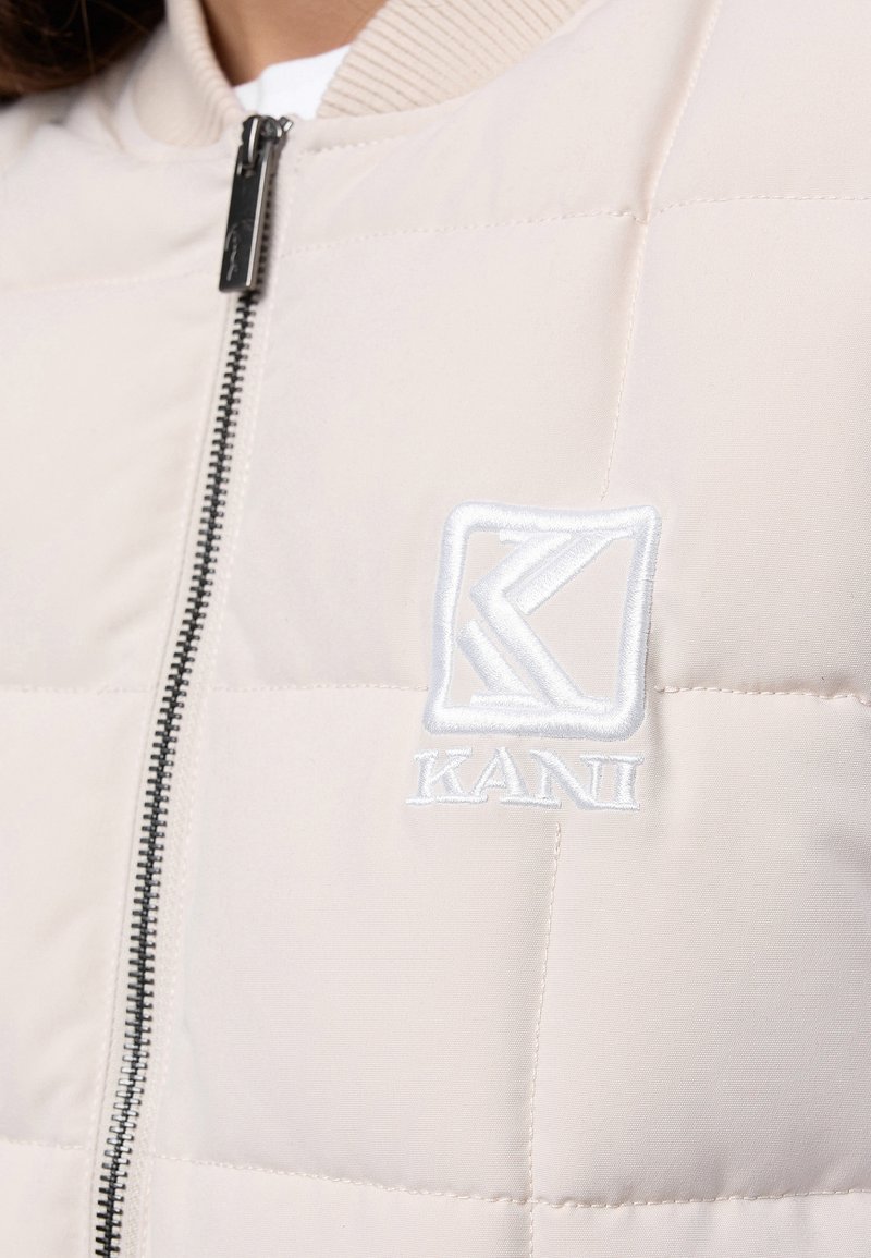 Beżowa pikowana kurtka z czarnym zamkiem i białym wyszywanym logo "KANI" na piersi.