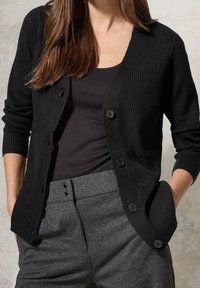 Schwarze gerippte Strickjacke mit V-Ausschnitt, Knopfverschluss und langen Ärmeln. Über einem schwarzen Oberteil getragen, kombiniert mit grauen strukturierten Hosen.