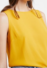 Bezramionkowy żółty top z gładkiego materiału, z prostym okrągłym dekoltem i luźnym krojem, z widocznymi detalami przeszycia.