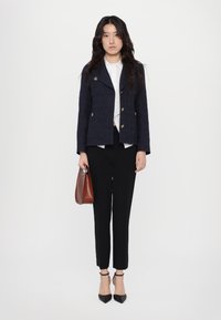 Navy teksturowana marynarka z złotymi guzikami, biała koszula pod spodem, czarne spodnie, czarne szpilki i brązowa torebka.