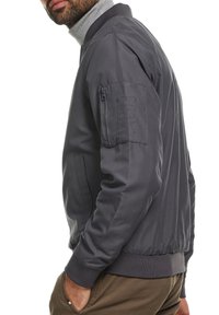 Grijze bomberjack met een ritsluiting, ribbelkraag en manchetten. Heeft een ritszak op de mouw en een glad, textuur afgewerkt oppervlak.