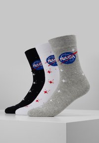 Trys poros paslaugų kojinių juodos, baltos ir pilkos spalvų. Kiekviena turi NASA logotipą ir raudonus bei baltus žvaigždžių raštus. Medžiaga yra medvilnės mišinys.