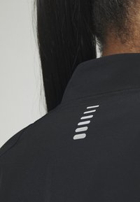 Gros plan du dos d’une personne portant une veste noire avec un motif de bande réfléchissante verticale sur le haut du dos sous le col.