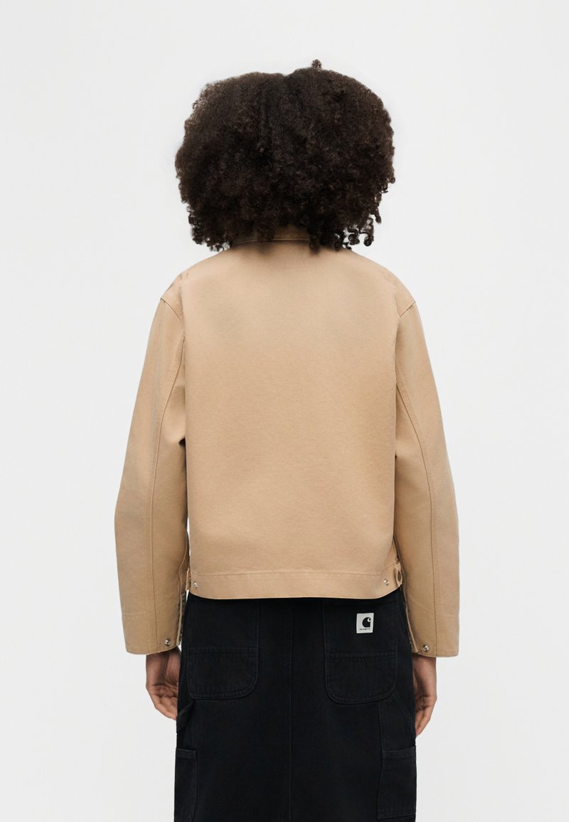 Person with curly hair wearing a beige jacket and black skirt standing facing away against a plain light background.