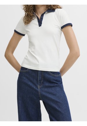 Frau trägt ein weißes Poloshirt mit marineblauem Kragen und Bündchen, kombiniert mit hoch taillierten blauen Jeans, steht mit den Händen am Rücken.