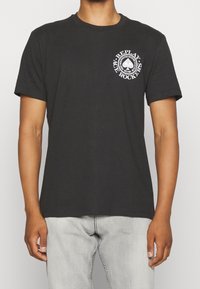 Osoba ubrana w czarną koszulkę z krótkim rękawem z białym logo "Replay Ace Rockers" na piersi oraz jasnoszare dżinsy.