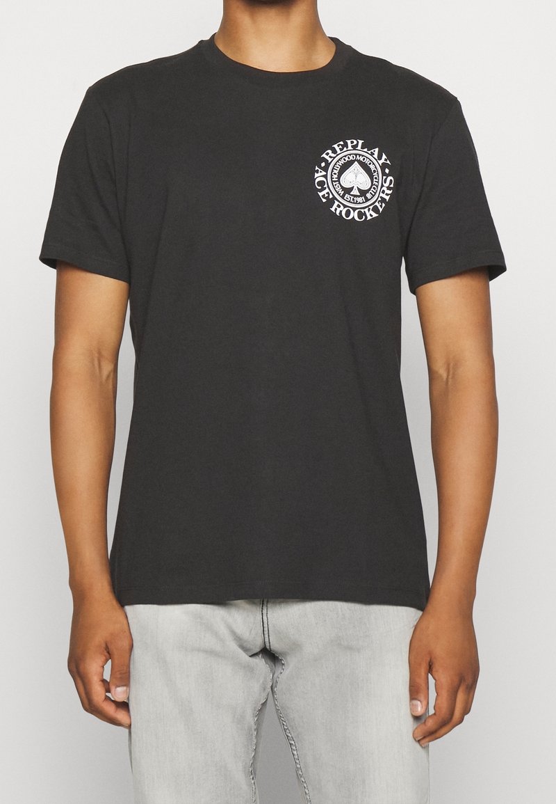 Osoba ubrana w czarną koszulkę z krótkim rękawem z białym logo "Replay Ace Rockers" na piersi oraz jasnoszare dżinsy.