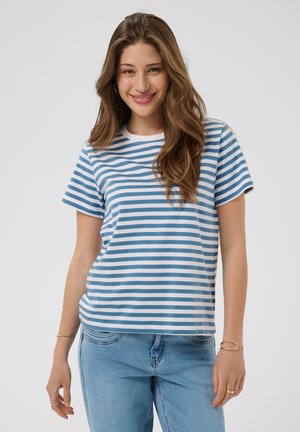 Ung kvinde smiler, iført en blå og hvid stribet kortærmet skjorte og lyseblå jeans, stående mod en ensfarvet baggrund.