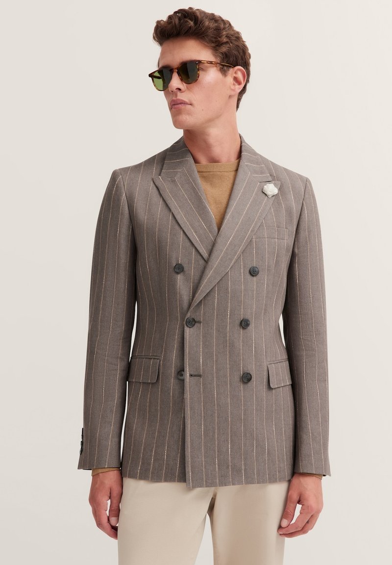 Grau gestreifter doppelreihiger Blazer mit schwarzen Knöpfen, Reverskragen und einer floralem Anstecker. Getragen über einem beigen Pullover, kombiniert mit hellen Hosen.