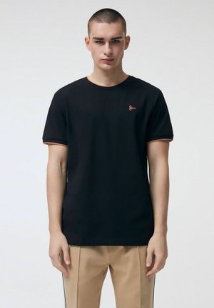 Młody mężczyzna o krótkich włosach, ubrany w czarną koszulkę z pomarańczowym wykończeniem i beżowe spodnie, stojący na tle jednolitego białego tła.