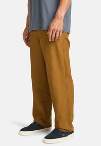 Pantaloni in cotone marrone con un design a gamba dritta, caratterizzati da cuciture laterali e abbinati a sneaker blu navy. La vestibilità è rilassata, adatta per un abbigliamento casual.
