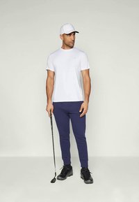 Moški, ki drži golf palico, nosi belo kratkodlako majico, mornarske hlače za šport, črne atletske čevlje in belo kapo.