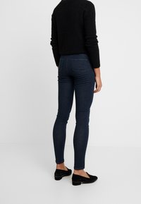 Smaljeans i mörkblå denim med medelhög midja. Bakifrån visas klassisk sömnad och två bakfickor. Stylet med en svart topp och platta skor.