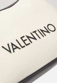 Canvas clutch i krämfärg med svart "VALENTINO"-text. Har en smidig läderkant och synliga sömmar längs kanterna.