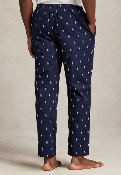 Άνδρας που φοράει ναυτικό μπλε παντελόνι πιτζάμας με λευκά λογότυπα αλόγων, στέκεται ξυπόλητος μπροστά σε απλό φόντο, με τα χέρια στις τσέπες.