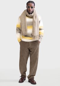 Pull en tricot rayé gris, jaune et blanc ; écharpe beige ; pantalon en velours côtelé marron ; chaussures marron à lacets. Textures de tissu lisses.