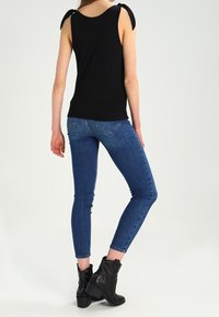 Vrouw in een zwarte mouwloze top met gestrikte schouderbanden, blauwe skinny jeans en zwarte enkelboots met studs, staand tegen een witte achtergrond.