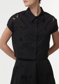 Black cropped shirt with short sleeves, featuring intricate black floral embroidery and a classic collar. Made of smooth, textured fabric.