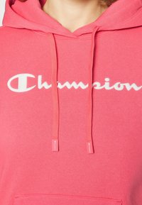 Růžová mikina s kapucí a bílým logem "Champion", se stahovacími šňůrkami s logem a strukturovanou tkaninou. Obsahuje přední klokaní kapsu.