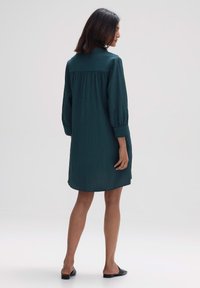 T türkisfarbene Knielange Kleid mit einem plissierten Muster, langen Ärmeln und einem Knopfkragen. Glattes Material mit einer lockeren Silhouette. Schwarze flache Schuhe.