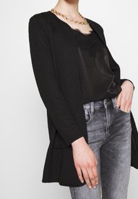 Chaqueta negra acolchada con frente abierto, combinada con una camisola de satén negra con detalles de encaje y jeans ajustados grises. Collar de cadena dorada visible.
