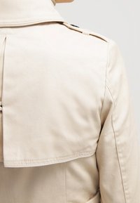 Trench-coat beige avec un design croisé, un pli dans le dos et des épaulettes. Tissu lisse, coupe ajustée et coutures nettes.