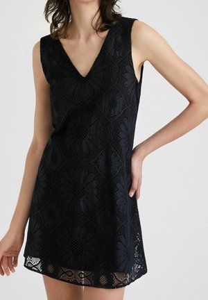 Robe en dentelle noire avec un décolleté en V, sans manches, ornée de motifs floraux et d'un ourlet festonné. La texture est ouverte et complexe.