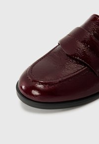 Maroon leather loafer with a glossy finish, featuring a wide strap across the vamp and contrasting black rubber sole. Stitching detail visible.