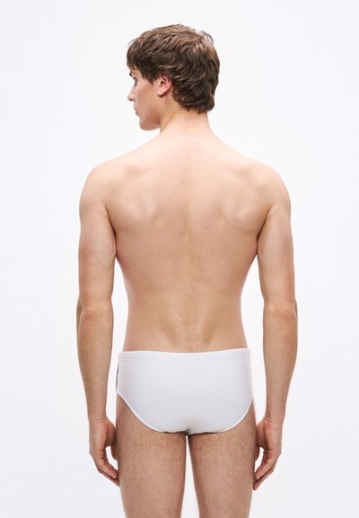 Weiße Badehose mit glatter Textur, figurbetontem Design und einfachem elastischem Bund. Der nackte Rücken des Models ist sichtbar und zeigt eine schlanke Statur.