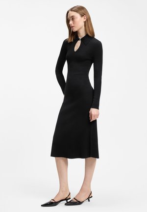 Kobieta ubrana w dopasowaną czarną sukienkę midi z długim rękawem i dekoltem typu keyhole oraz czarne niskie buty typu slingback, stoi bokiem.