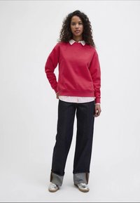 Sweatshirt vermelho, camisa de colarinho listrada em branco e rosa por baixo, calças de ganga escuras, sapatos prateados com detalhes distintos, cabelo encaracolado.
