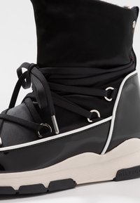 Czarny botek za kostkę z połyskującym noskiem z lakierowanej skóry, materiałową cholewką, wiązany na sznurówki, z srebrnymi przelotkami i grubą gumową podeszwą z białymi akcentami.