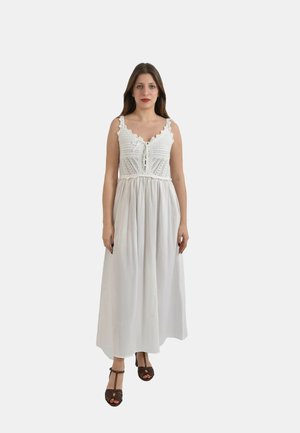 Donna in piedi che indossa un vestito bianco senza maniche con corpetto allacciato in pizzo lavorato a uncinetto e gonna fluente, abbinato a tacchi marroni aperti.
