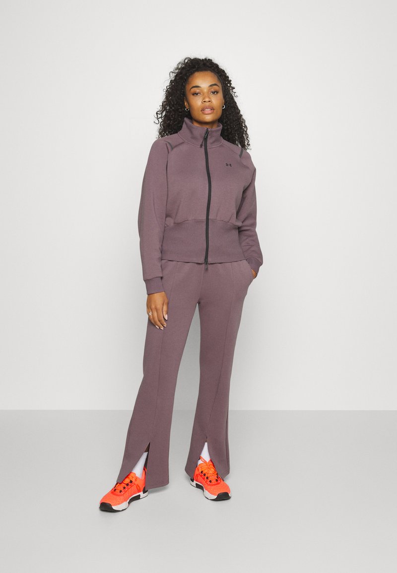 Lila Sportjacke mit Reißverschluss und passender Hose mit seitlichen Schlitzen. Helle orangefarbene Sneaker runden das Outfit ab. Glatte Textur, sportliches Design.