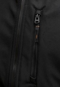 Nærbilde av et svart stoff med to vertikale glidelåser, hvorav den ene har en glideklaff med oransje søm.