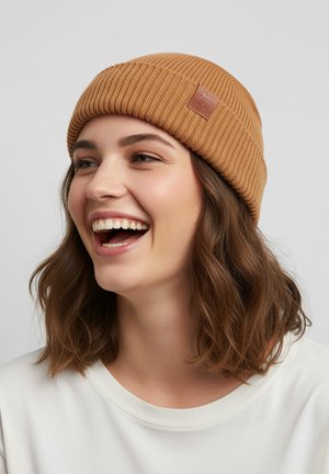 Braune gerippte Strickmütze mit einem Lederetikett, getragen von einer Person mit welligem Haar. Weiche Textur, lässiges Design, eng anliegende Passform.