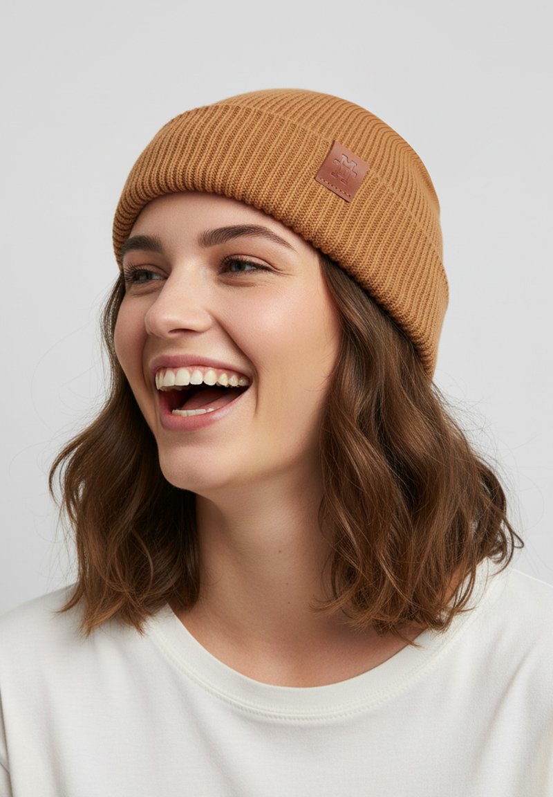 Brown ribbed knit beanie with a leather label, worn by a person with wavy hair. Soft texture, casual design, snug fit.