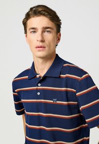 Marinblå polo med horisontella röd- och krämfärgade ränder, blandning av bomull, tre-knapps knäppning och en liten logotyp på vänster bröst.